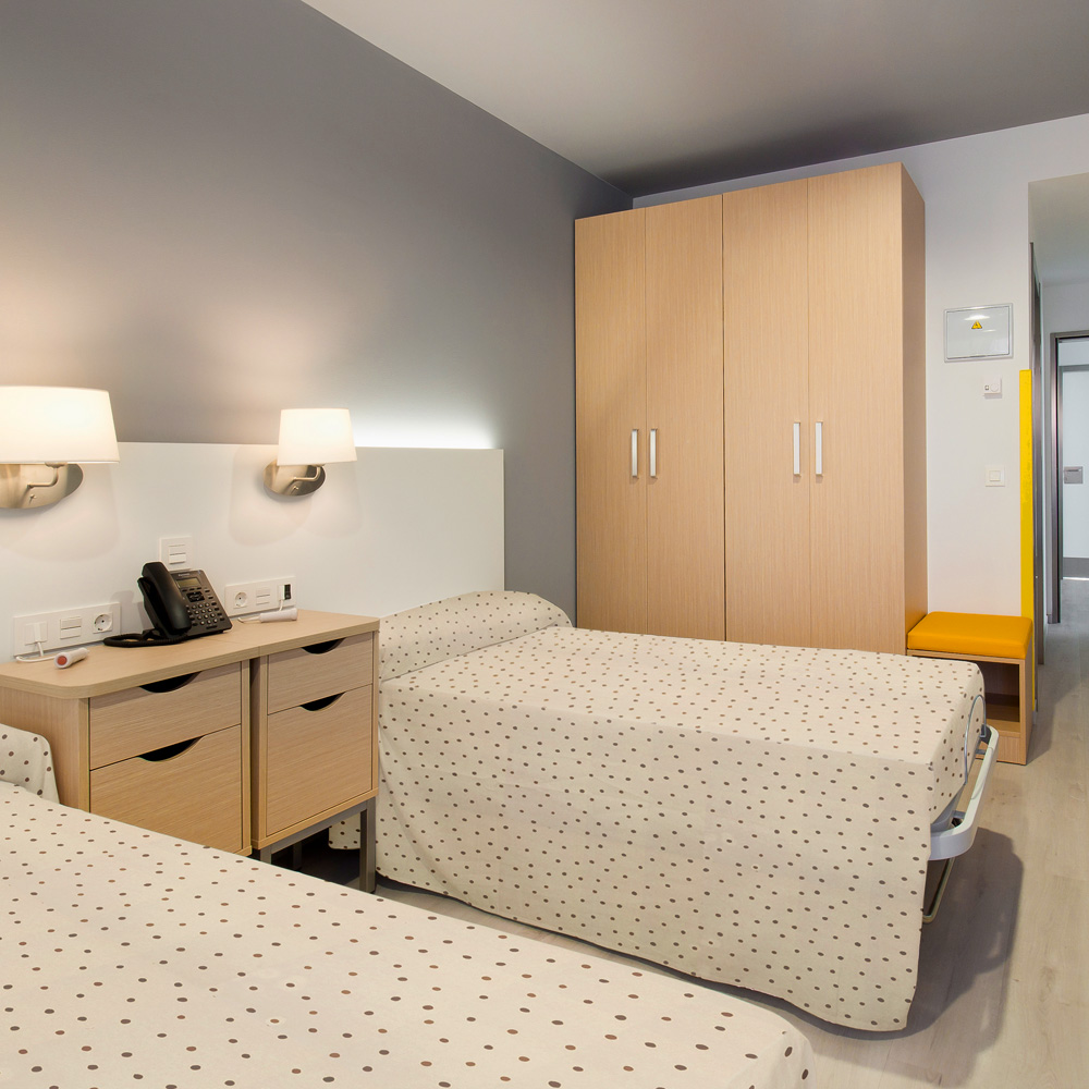 Vista de una habitación doble de la residencia de mayores CleceVitam Altos de Parquesol con dos camas, mesillas de noche y armarios